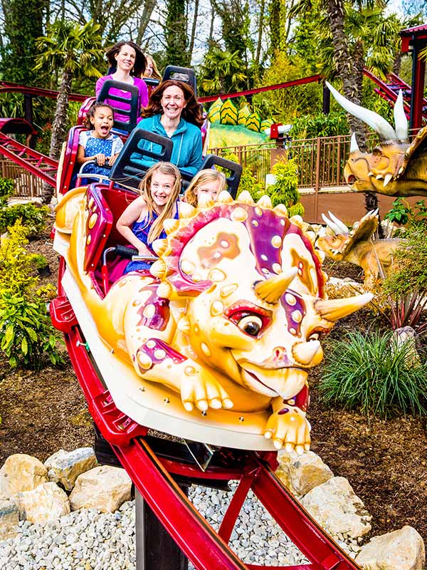 Children and parents riding Dino Chase roller coaster at Paultons Park, smiling in triceratops themed train car