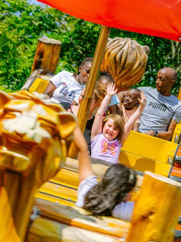 Children smiling with hands in the air on Kontiki at Paultons Park surrounded by green trees on a sunny day