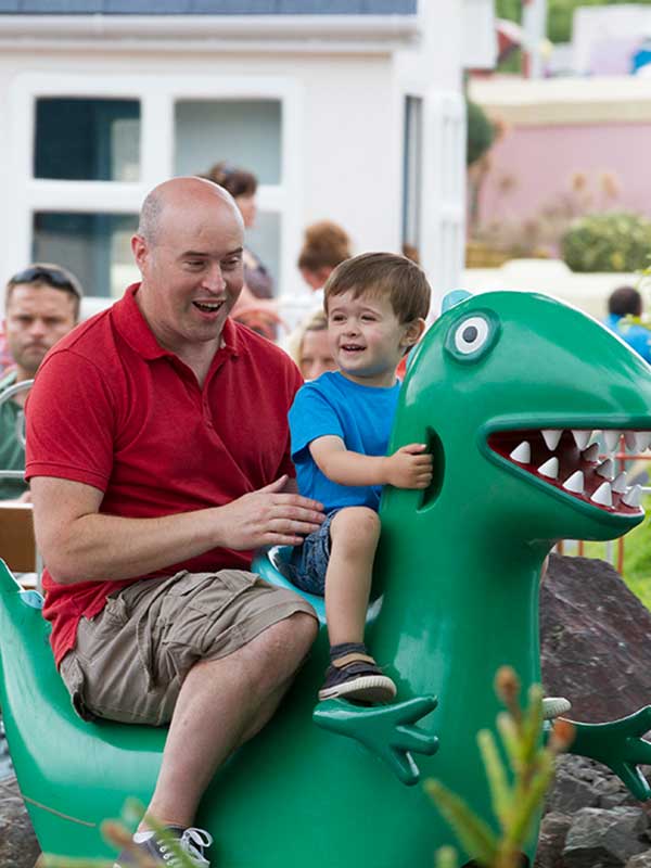 Father and young child riding a green dinosaur car on George’s Dinosaur Adventure in Peppa Pig World at Paultons Park