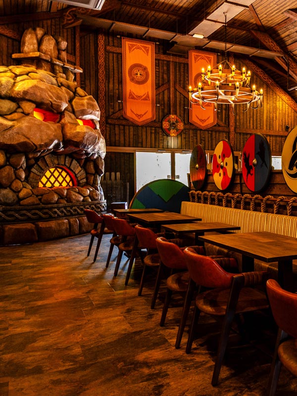 Viking-themed interior of the Feasting Hall in Valgard at Paultons Park, with wooden tables, shields and a glowing stone fireplace.