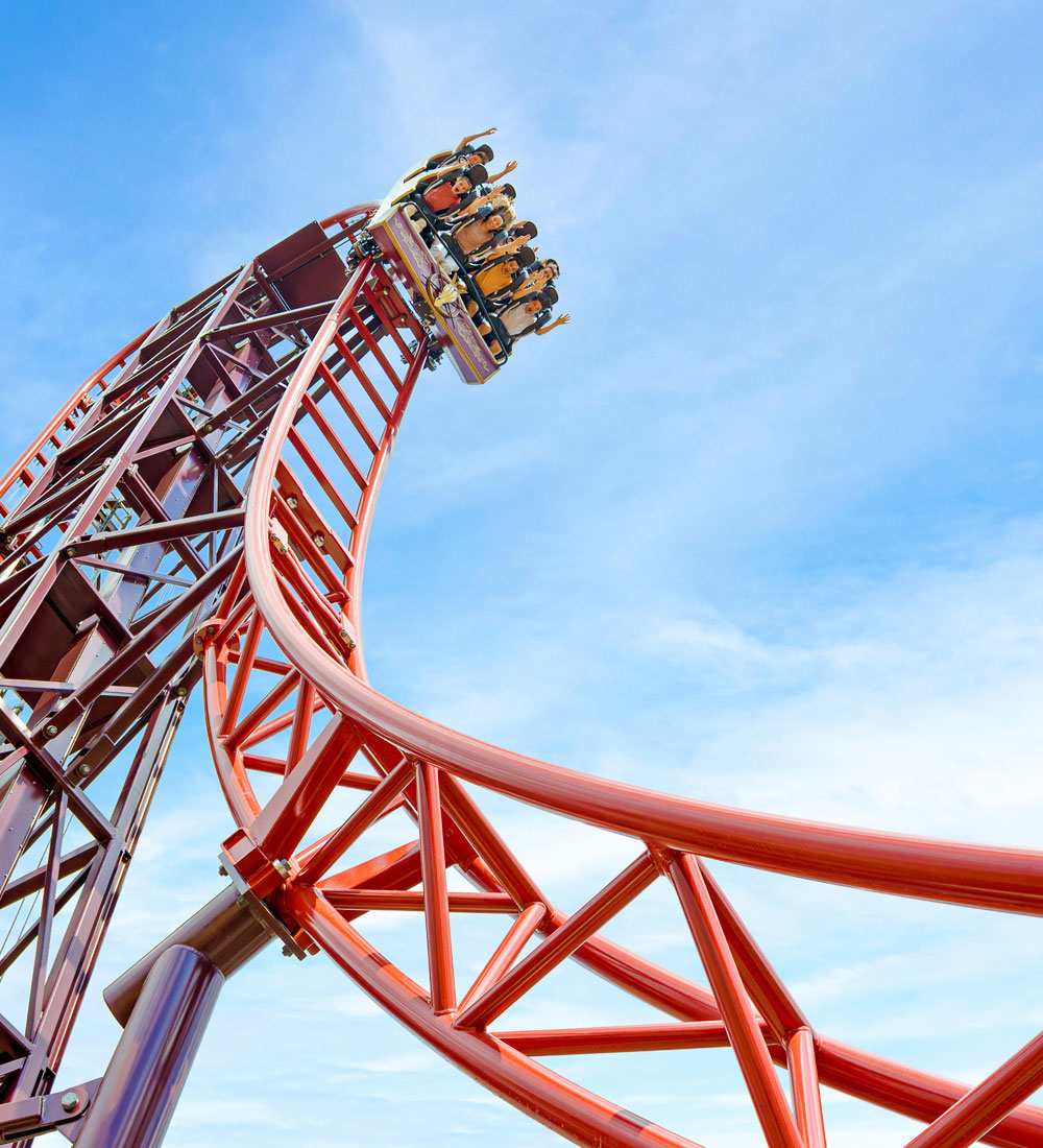 Riders crest the towering red track of Drakon, the Euro-Fighter rollercoaster in Valgard at Paultons Park, against a bright blue sky